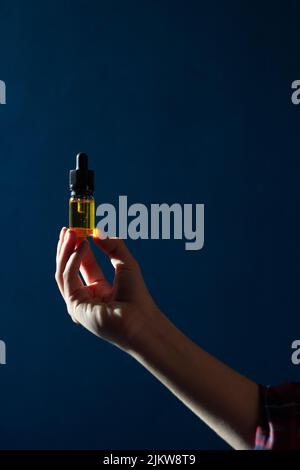 In der Hand einer Frau befindet sich eine Glasflasche mit einer Pipette, die mit cbd-Öl gefüllt ist. Auf einem dunkelblauen Hintergrund Stockfoto