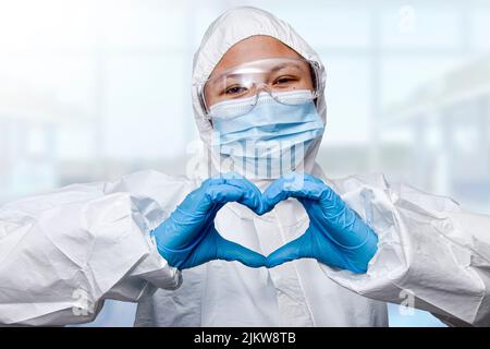 Eine Apothekerin mit einer schützenden medizinischen Maske und einer Brille, die mit den Händen eine Herzform formt Stockfoto