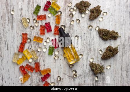 Eine Glasflasche mit gelbem CBD-Cannabisöl liegt auf einem Holztisch, umgeben von trockenen medizinischen Marihuanaknospen, cbd-Extraktkapseln und buntem cbd-Kaugummi Stockfoto