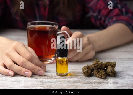 Die Hände der Frauen halten eine Tasse Tee. Im Vordergrund eine Glasflasche mit einer Pipette mit Cannabis cbd-Öl, trockene Marihuanaknospen liegen in der Nähe Stockfoto