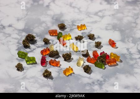 Mehrere trockene Knospen von medizinischem Marihuana liegen zufällig unter Gummibären verschiedener Farben mit der Zugabe von cbd-Öl auf einem Marmortisch Stockfoto