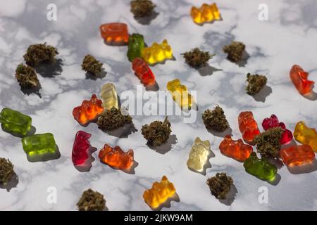 Mehrere trockene Knospen von medizinischem Marihuana liegen zufällig unter Gummibären verschiedener Farben mit der Zugabe von cbd-Öl auf einem Marmortisch Stockfoto