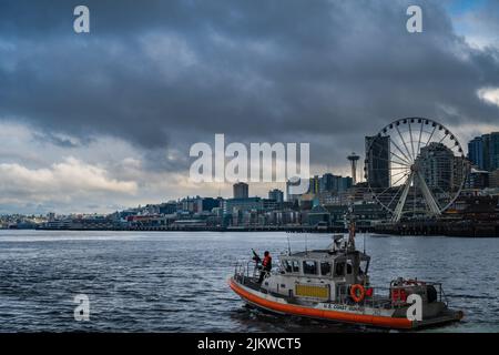 DIE HAFENPROMENADE VON SEATTLE MIT DER KÜSTENWACHE, DIE ELLIOTT BAY PATROUILLIERT, MIT DEM GROSSEN RAD UND DER WELTRAUMNADEL IM HINTERGRUND Stockfoto