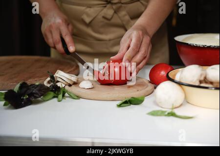 Die Hände des Küchenchefs schneiden Tomaten auf einem Schneidebrett mit in Scheiben geschnittenen Champignons und kulinarischen aromatischen Kräutern auf dem Küchentisch Stockfoto