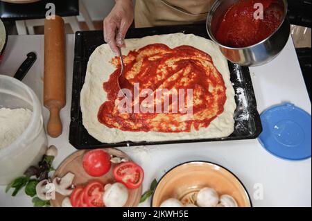Draufsicht. Prozess der Zubereitung von Pizza. Der Koch gießt Tomatensauce über den gerollten Teig. Frische Zutaten auf dem Küchentisch Stockfoto