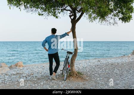 Ein junger Mann mit einem Fahrrad, das sich auf den Baum lehnt und auf das Mittelmeer blickt. Stockfoto