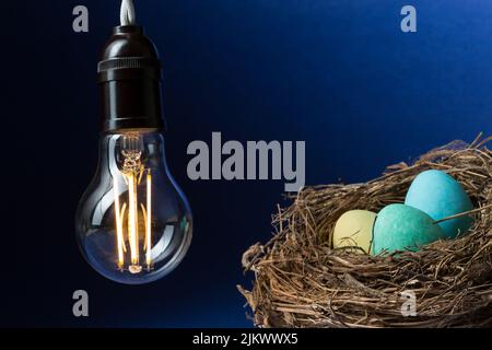 Nahaufnahme einer beleuchteten LED-Lampe, die drei farbige Eier in einem Vogelnest auf schwarzem und blauem Hintergrund erhitzt. Stockfoto