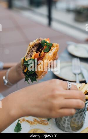 Frau, die einen halb gefressenen Burger in der einen Hand hält und Pommes mit der anderen Hand isst Stockfoto