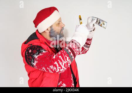 Neujahrs- und Weihnachtskonzept. Der Weihnachtsmann untersucht Geld durch eine Lupe. Stockfoto