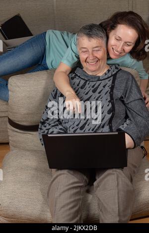 Lächelnd zwei Frauen, die auf den Laptop-Bildschirm schauen, die Finanzen überprüfen und zu Hause am Sofa sitzen. Glückliche Mutter und Erwachsene Tochter machen Online-Shopping Stockfoto