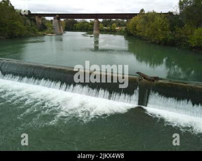 Ein Blick auf einen Damm auf einem Fluss, der durch seine mit Grün bedeckten Ufer fließt Stockfoto