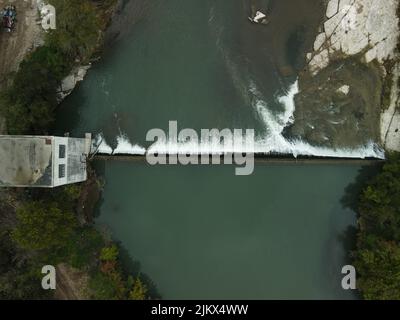 Ein Luftbild von oben auf einem Damm auf einem Fluss, der durch seine mit viel Grün und einem Gebäude bedeckten Ufer fließt Stockfoto