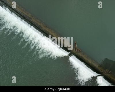 Ein Luftbild von oben auf einem Damm auf einem Fluss, der durch seine mit Grün bedeckten Ufer fließt Stockfoto