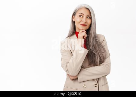 Bild einer eleganten asiatischen Senior-Frau im Anzug, lächelnd und fasziniert, nachdenklicher Gesichtsausdruck, der auf weißem Hintergrund steht Stockfoto