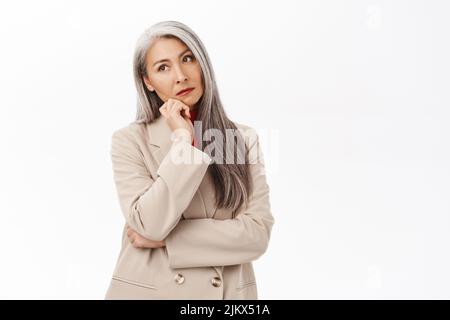 Porträt einer koreanischen Geschäftsfrau, Frau mit grauen Haaren denkend, Anzug, lächelnd fasziniert, stehend auf weißem Hintergrund Stockfoto