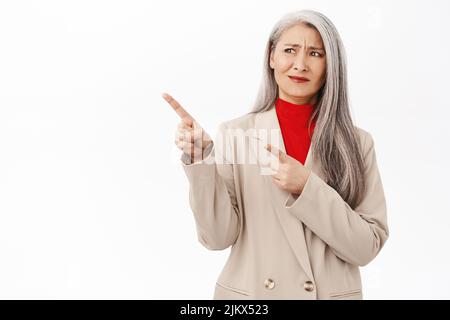 Asiatische Geschäftsfrau, Unternehmerin zeigt mit enttäuschtem Gesicht nach links und steht aufgeregt vor weißem Hintergrund Stockfoto