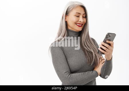 Stilvolle asiatische ältere Frau, schöne Dame hält Handy, posiert vor weißem Hintergrund mit glücklich lächelnd Gesichtsausdruck Stockfoto