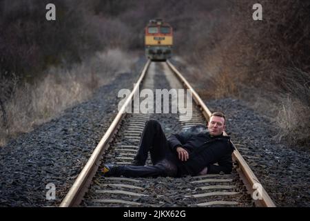Junger Mann, der auf der Eisenbahnstrecke liegt und eine Zigarette raucht, während ein Zug im Hintergrund kommt Stockfoto