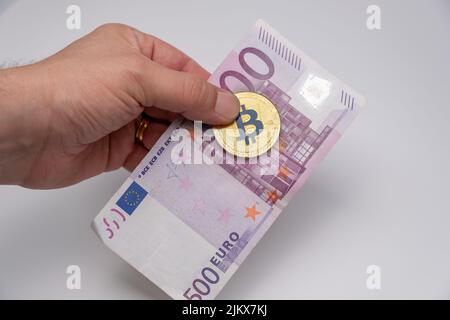 Männliche Hand hält Geld. Goldbitcoin und 500 Euro Banknote. Goldene BTC-Münze der Kryptowährung und eine fünfhundert-Euro-Banknote auf Männerfi Stockfoto