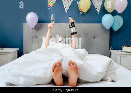Frau liegt auf dem Bett mit erhobenen Händen halten Flasche und Glas Champagner in dekorierten Schlafzimmer.Pläne für Geburtstag. Zeit für sich selbst. Feiern Stockfoto