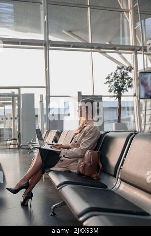 Kaukasische Dame, die auf dem Laptop tippt und in der Wartehalle am Flughafen sitzt Stockfoto