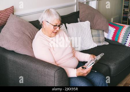 Alte ältere Frau, 80s Erwachsene, Großmutter, die Kreuzworträtsel in ihrem Wohnzimmer macht, das auf dem Sofa sitzt. Ruhestandhobby für Trainingsgedächtnis. Alzhe Stockfoto