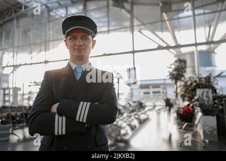 Glücklicher erwachsener männlicher Pilot im Hut, der vor der Kamera im Terminal posiert Stockfoto