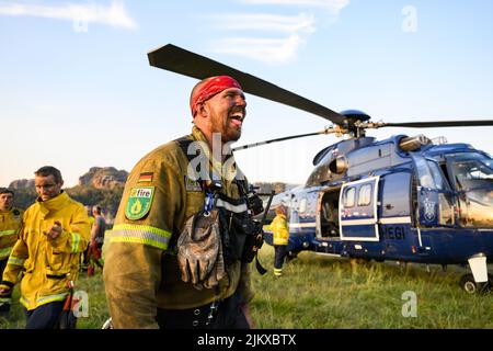 Bad Schandau, Deutschland. 03. August 2022. Freiwillige Feuerwehrleute des Internationalen Katastrophenschutzes Deutschland '@Fire' stehen nach dem Einsatz gegen die Waldbrände im Nationalpark Sächsische Schweiz vor einem Hubschrauber der Bundespolizei auf einer Wiese. Der Internationale Katastrophenschutz Deutschland e. V. ist eine gemeinnützige, nichtstaatliche Hilfsorganisation, die im Falle von Naturkatastrophen internationale Hilfe leistet. Kredit: Robert Michael/dpa/Alamy Live Nachrichten Stockfoto