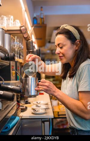 Lächelnde Kellnerin in der weiblichen Cafeteria, die Kaffee in einer Kaffeemaschine zubereitet, kovidische Einschränkungen werden aufgehoben und die obligatorische Verwendung von Gesichtsmasken wird entfernt Stockfoto