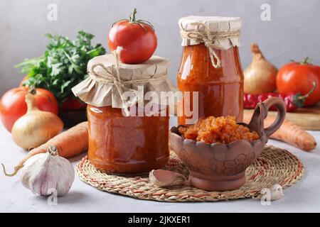 Hausgemachter Gemüsekaviar aus Karotten, Paprika, Knoblauch und Tomaten in Schüssel und Gläsern auf grauem Hintergrund Stockfoto