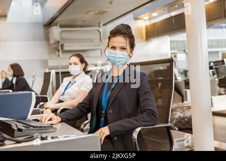 Frau in der Fluggesellschaft, die Gesichtsmaske trägt, während sie am Check-in-Schalter der Fluggesellschaft am Flughafen arbeitet Stockfoto