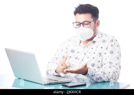 ein indischer Mann trägt eine Maske und desinfiziert die Hand, während er in einem Büro einen Laptop benutzt Stockfoto