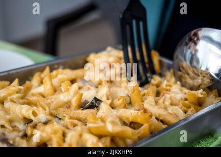 Bild von Tablett mit Macaroni Pasta mit geschmolzenem Käse und Gemüse, das während einer Mahlzeit mit Paddel und großem Löffel am Tisch serviert wird Stockfoto