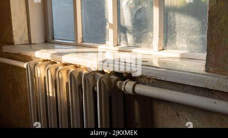 Altes Vintage-Haus im Sonnenuntergang Licht. Gusseiserner Heizkörper und Holzfenster. Weiße Fensterbank mit schäbiger Farbe auf dem Hintergrund der Wände ohne Tapete. Retro-Hintergrund Stockfoto