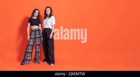 Zwei Frauen, Tochter und Mutter, in schwarzen Hosenanzügen, stehen vor orangefarbenem Hintergrund, schauen auf die Kamera und lächeln. Negatives Leerzeichen rechts. Stockfoto