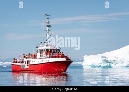 Leuchtend rotes Fischerschiff, das am 17. Juli 2022 Touristen zwischen großen Eisbergen im Ilulissat-Eisfjord, Grönland, transportiert Stockfoto