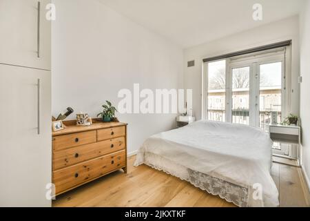 Komfortables Bett mit gepunkteter Federdecke in der Nähe von Kleiderschrank und Tisch mit Stuhl im minimalistischen Stil im modernen Apartment Stockfoto