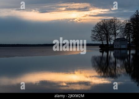 Eine schöne Aufnahme von großen, blattlosen Bäumen und ländlichem Haus am Seeufer mit sichtbaren Reflexionen auf dem Wasser gegen den Abendhimmel bei Sonnenuntergang Stockfoto