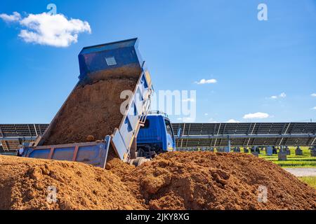 Ein blaues sechsrädriges Fahrzeug gießt orangenen Boden auf den Boden eines Industriegeldes, um einen durch Erosionswasser beschädigten Brunnen an der Solarstromquelle zu füllen Stockfoto