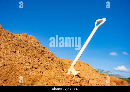 Eine weiße Schaufel wurde auf einen Haufen orangefarbener Erde mit einem blauen Himmel und ein paar Wolken eingesetzt. Bauarbeiten in Industrieanlagen. Stockfoto