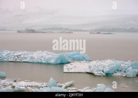 Wie türkisfarbene Eisberge schweben sie auf dem Fjallsarlon-Gletschersee in Island. Entfernte Berge im Hintergrund sichtbar. Stockfoto