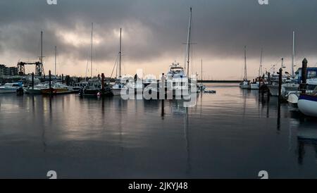 Ein grauer Sommerabend-Regenschauer an einem Sportboothafen und die Spiegelungen der Boote im Meerwasser. Stockfoto