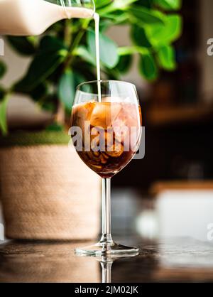 An einem Sommertag wird Milch in ein Glas Eiskaffee am Fenster gegossen. Stockfoto