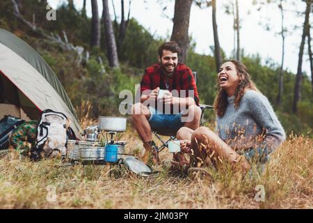 Glückliches junges Paar, das vor ihrem Zelt lachend sitzt. Fröhliches junges Paar, das sich auf einem Campingurlaub amüsieren kann. Stockfoto