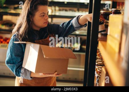 Supermarktmitarbeiter mit Down-Syndrom stocken Lebensmittel in die Regale. Ermächtigte Frau mit einer intellektuellen Behinderung, die als A s arbeitet Stockfoto