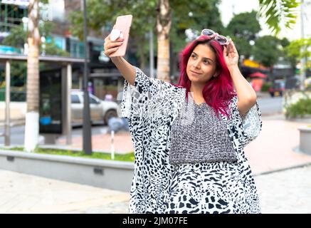 Eine junge hispanische Hündin mit rosa Haaren in einem gemusterten Outfit, die für ein Selfie posiert Stockfoto