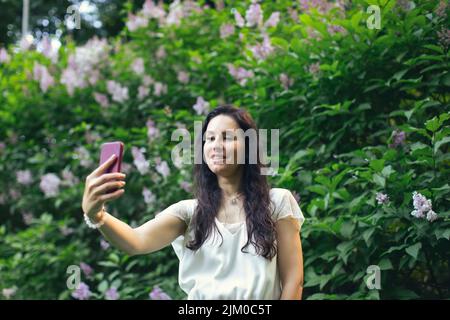 Attraktive Frau, die Selfies mit einem Busch blühender Flieder macht. Frühling und Sommer, saisonale Fotos. Vorderansicht. Stockfoto