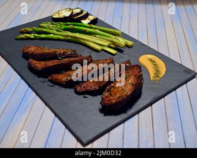 Nahaufnahme eines in Scheiben geschnittenen veganen Fleisches, serviert mit Spargel und gegrillten Auberginen und einer gelben Sauce Stockfoto