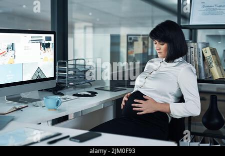 Nur mein Baby und ich im Büro. Eine attraktive Schwangere, die allein im Büro sitzt und ihren Magen hält. Stockfoto