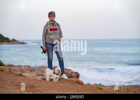 Eine junge Hündin mit ihrem maltesischen bichon, die eine tolle Zeit am Strand von Oropesa del mar in Castellon Spanien hat Stockfoto